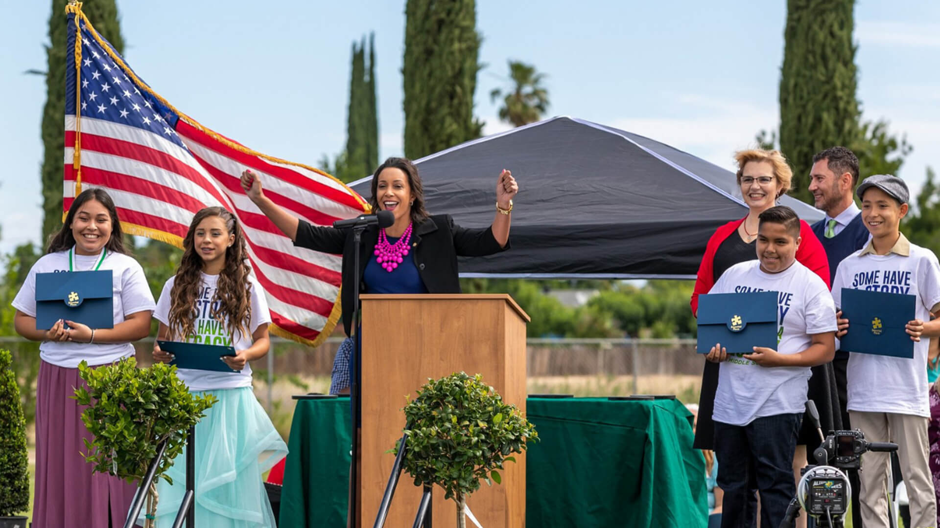 United Way Fresno and Madera Counties President and CEO Lindsay Fox cheers behind a podium on stage at an outdoor event. Smiling children stand on each side of her.